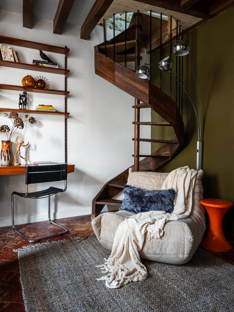 Coin lecture avec fauteuil en velours côtelé, escalier hélicoïdal en bois et tomettes anciennes dans une maison rénovée en Centre-Val de Loire