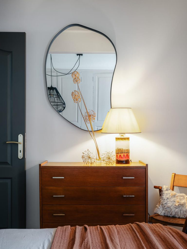 Commode vintage en bois massif avec miroir organique, lampe en céramique années 70 et fleurs séchées dans une chambre d'hôtes