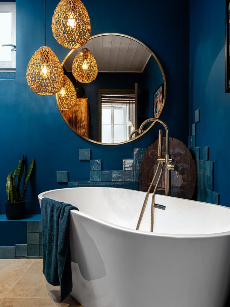 Salle de bains bleu profond avec trio de suspensions en osier, baignoire ilot blanche, miroir rond doré et zellige artisanal
